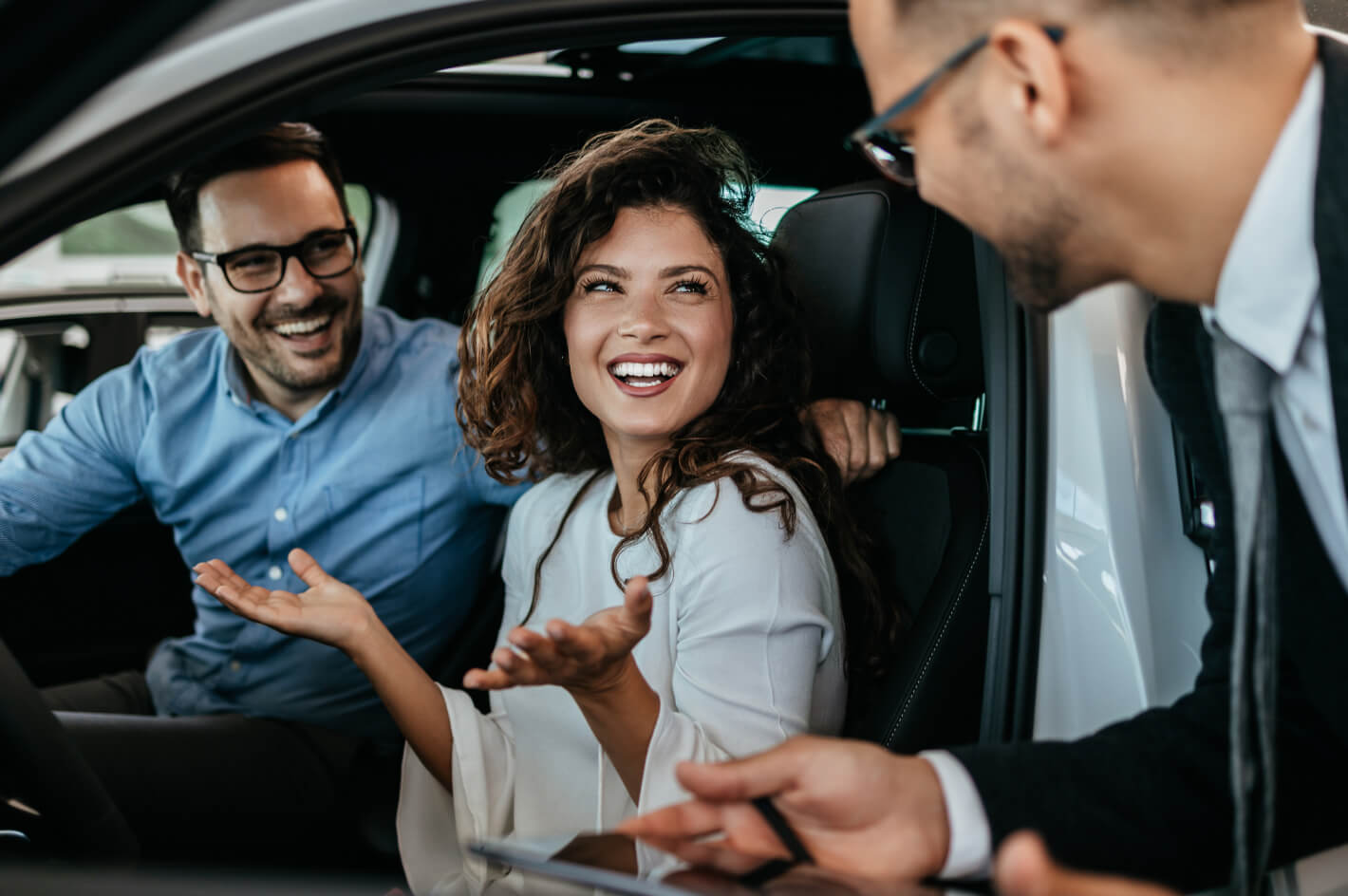 A sales associate leans towards an open car door as a couple smiles while sitting in the driver and front passenger seats.