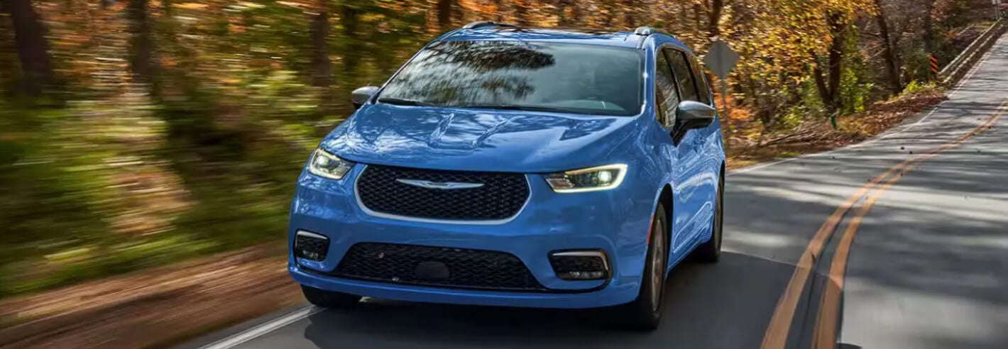 Front view of a blue Chrysler Pacifica driving down a tree-lined road.